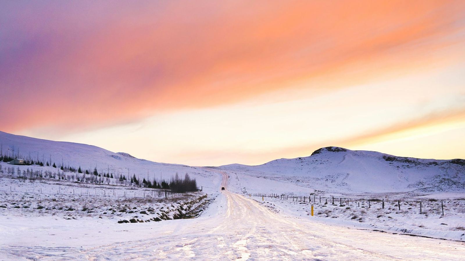 A snow-covered road through white hills in Iceland under a pink and orange winter sky